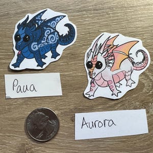 K&ouml;nnte beinhalten: Zwei gestanzte Aufkleber mit Cartoon-Drachen mit gro&szlig;en schwarzen Augen. Ein Drache ist blau mit dunkelblauen Wirbelmustern und mit 'Paua' beschriftet. Der andere ist wei&szlig; mit rosa und orangefarbenen Fl&uuml;geln und mit 'Aurora' beschriftet.