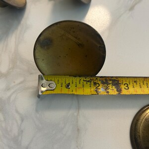 Könnte beinhalten: Nahaufnahme von antiken Messing-Schrankknöpfen. Ein Knopf wird mit einem Maßband gemessen und zeigt einen Durchmesser von etwa 6,35 cm. Die Knöpfe haben ein rundes, leicht gewölbtes Design mit einem angelaufenen Finish, was auf Alter und Nutzung hindeutet.