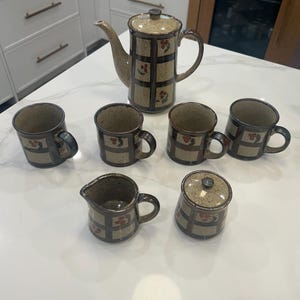 May include: A ceramic tea set comprising a teapot, six mugs, a creamer jug, and a sugar bowl. The set has a beige base with a brown grid design and red floral details. The teapot has a long spout and a matching lid.