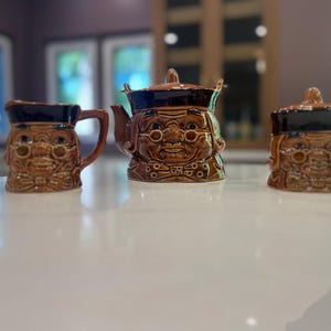 Peut inclure: Un service à thé en céramique marron comprenant une théière, un pot à crème et un sucrier. Chaque pièce est en forme de personne âgée souriante avec des lunettes et un chapeau. L'ensemble a un glaçage marron foncé sur le dessus et plus clair sur le visage.