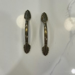 May include: Two antique-style cabinet handles. Each handle is a bronze colour with a vertical, ribbed design and decorative, fan-shaped ends. The handles are set against a white marble-patterned surface.