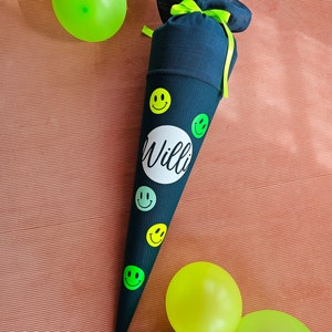 Könnte beinhalten: Ein schwarzer Schultüte mit einem grünen Band und grünen Smiley-Aufklebern. Die Tüte hat einen weißen Kreis mit dem Namen "Willi" in Schwarz.