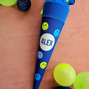 Può includere: Un cono scolastico blu e giallo con un cerchio bianco che dice "ALEX" e faccine sorridenti. Il cono è decorato con un fiocco di stoffa blu e bianco.