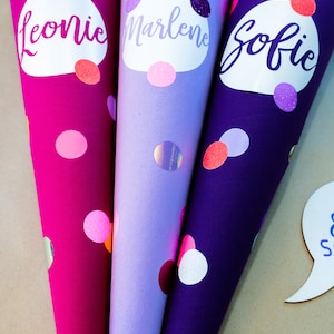 Puede incluir: Tres conos escolares coloridos en tonos rosa, lila y morado, cada uno adornado con un nombre y lunares. Los nombres "Leonie", "Marlene" y "Sofie" est&aacute;n escritos en cursiva sobre &oacute;valos blancos. Los conos est&aacute;n decorados con purpurina y detalles hologr&aacute;ficos.