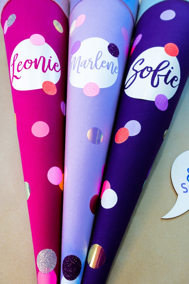 Puede incluir: Tres conos escolares coloridos en tonos rosa, lila y morado, cada uno adornado con un nombre y lunares. Los nombres "Leonie", "Marlene" y "Sofie" est&aacute;n escritos en cursiva sobre &oacute;valos blancos. Los conos est&aacute;n decorados con purpurina y detalles hologr&aacute;ficos.