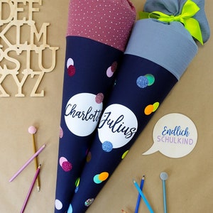 Puede incluir: Dos conos escolares azul marino con confeti colorido y nombres personalizados. Uno tiene una parte superior burdeos con lunares, el otro una parte superior gris. El texto "Endlich SCHULKIND" est&aacute; en una burbuja. Hay l&aacute;pices esparcidos.