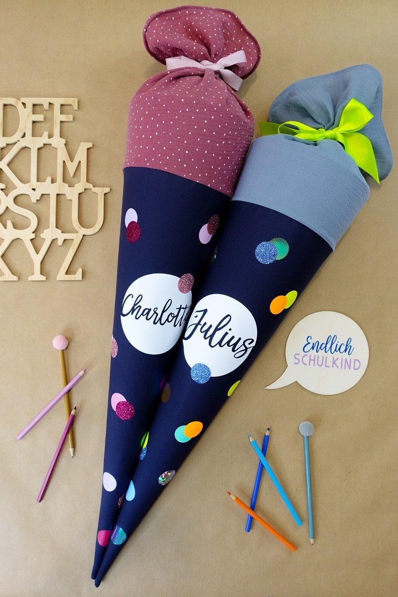 Puede incluir: Dos conos escolares azul marino con confeti colorido y nombres personalizados. Uno tiene una parte superior burdeos con lunares, el otro una parte superior gris. El texto "Endlich SCHULKIND" est&aacute; en una burbuja. Hay l&aacute;pices esparcidos.