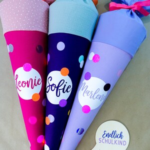 Puede incluir: Tres coloridos conos escolares con lunares y nombres personalizados. Las bolsas son en tonos rosa, morado y azul, con tapas de tela a juego atadas con cintas. El texto "Endlich SCHULKIND" est&aacute; en un bocadillo.