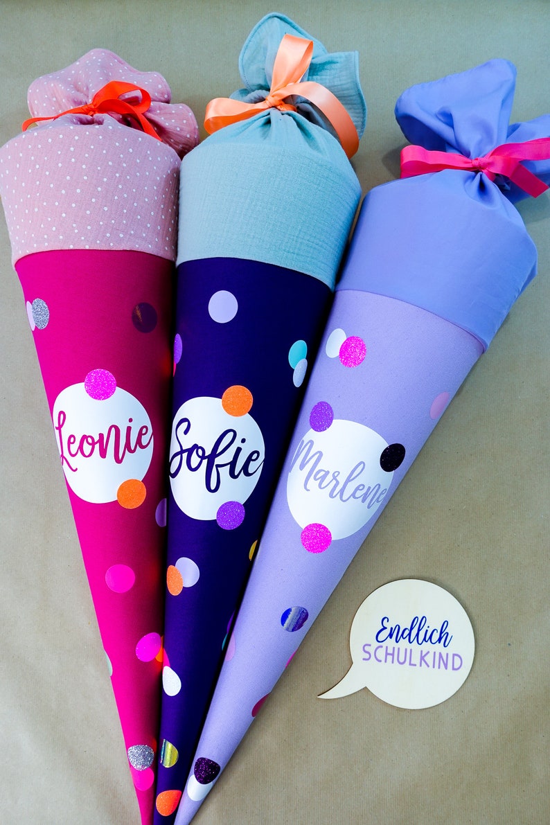 Puede incluir: Tres coloridos conos escolares con lunares y nombres personalizados. Las bolsas son en tonos rosa, morado y azul, con tapas de tela a juego atadas con cintas. El texto "Endlich SCHULKIND" est&aacute; en un bocadillo.