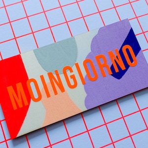 以下が含まれることがあります： 鮮やかなオレンジ色の文字で「MOINGIORNO」と書かれた長方形の木製サイン。赤、桃色、グレー、紫、青の形を使ったカラフルな抽象的なデザイン。背景は水色の格子模様。