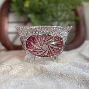 May include: A clear and cranberry red glass bowl featuring a diamond-cut pattern on the upper rim and a central oval panel with a starburst design. The bowl is supported by three small glass feet and has a scalloped edge along the top rim.