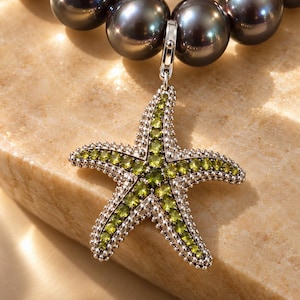 May include: A necklace featuring a strand of dark grey, spherical pearls and a silver-toned starfish pendant. The starfish is encrusted with small, vibrant green gemstones along its five arms, attached to the pearl strand by a silver clasp.