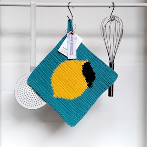 Könnte beinhalten: Ein türkisfarbener, gehäkelter Topflappen mit einem gelben Zitronenmotiv. Der Topflappen hängt an einem Metallhaken. Das Etikett auf dem Topflappen lautet "COOKIES INTERNATIONAL".