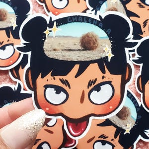 Può includere: Un adesivo con un personaggio dei cartoni animati con i capelli neri e una scena del deserto nella sua testa. L'adesivo ha il testo "CHALENA.A" e stelle scintillanti. Il personaggio ha un'espressione sorpresa.