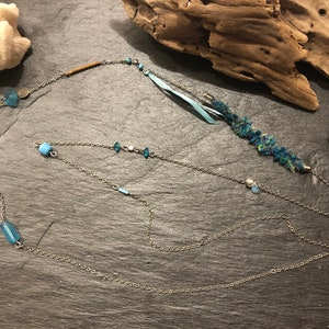Peut inclure: Un collier à chaîne argentée à plusieurs rangs avec des perles de verre bleues et un pompon en tissu bleu. Le collier est sur une surface gris foncé.