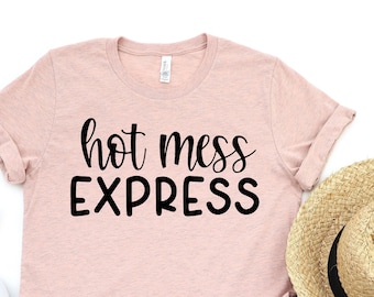 May include: A light pink t-shirt with the words "hot mess EXPRESS" in black script. The shirt is paired with denim shorts, a straw hat, white sneakers, and sunglasses.
