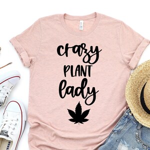Può includere: Una maglietta rosa con il testo "crazy plant lady" in corsivo nero e un grafico di foglia di marijuana nera.