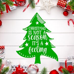 以下が含まれることがあります： 白い木の背景に、「Christmas is not a season it's a feeling」というテキストが入った緑色のクリスマスツリーのグラフィック。画像は、包装されたギフト、オーナメント、緑など、クリスマスの装飾で囲まれています。