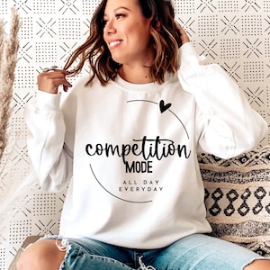 Pode incluir: Camisola branca com texto preto que diz "competition mode all day everyday" num círculo com um pequeno coração acima do texto.
