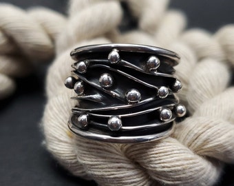 May include: A wide silver ring with a textured, wavy design and small silver beads. The ring is set against a white rope background.