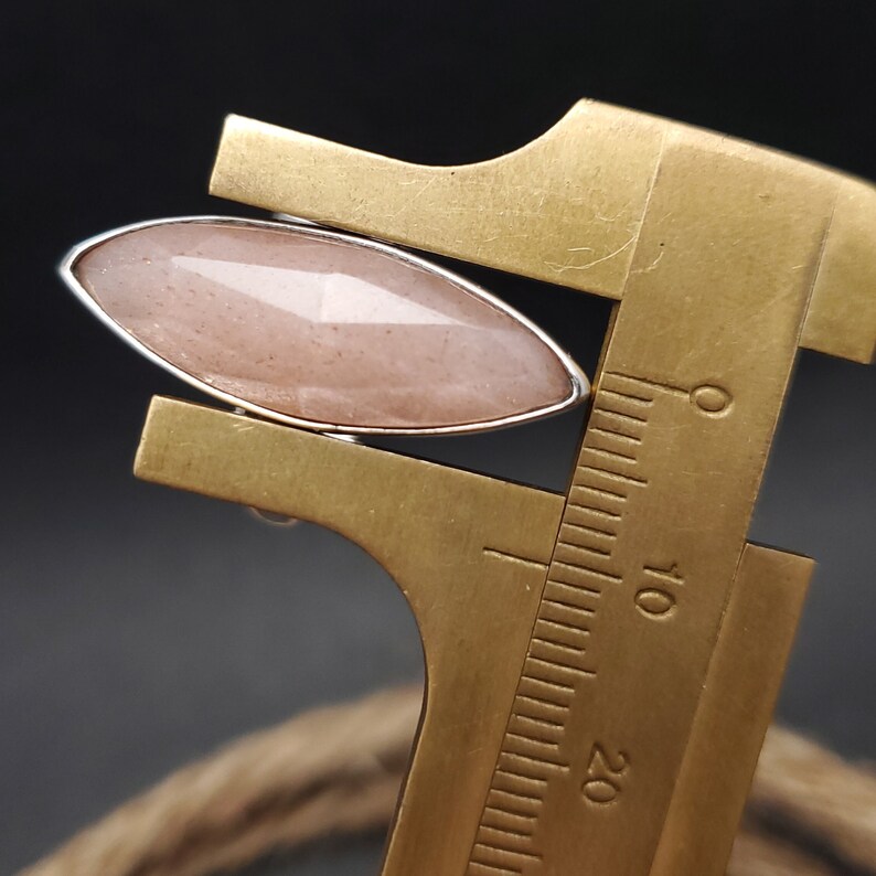 May include: A marquise-cut, light pink gemstone set in a silver-colored metal ring. The gemstone is faceted, reflecting light. A brass measuring tool is positioned near the ring, providing scale. The background is dark, with a glimpse of a rope.