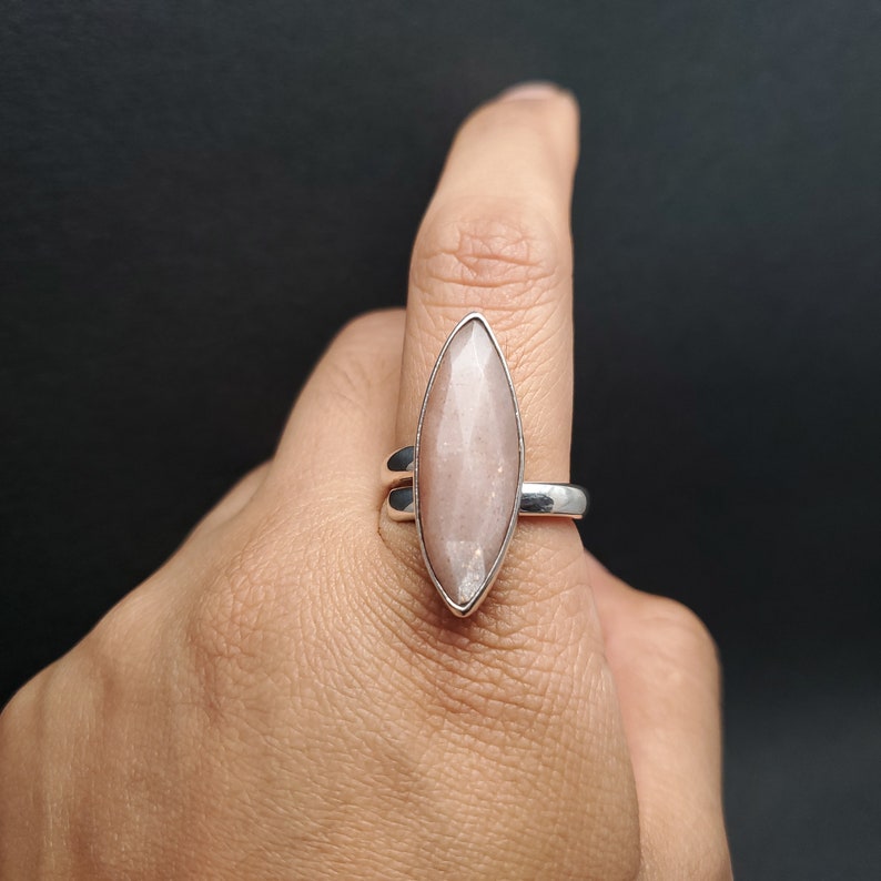 May include: A silver ring featuring a large, light pink, marquise-cut gemstone. The gemstone is set in a bezel setting and has a faceted surface. The ring has a simple, elegant design, suitable for everyday wear or special occasions.