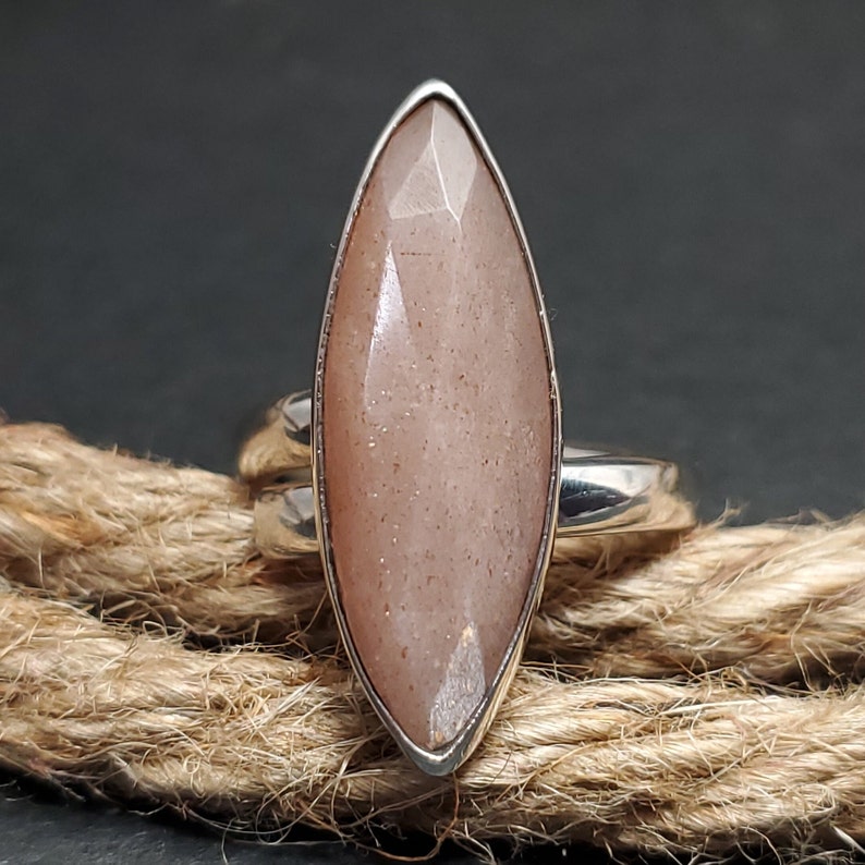 May include: A silver ring featuring a large, elongated, faceted, peach-colored gemstone. The gemstone is set in a silver bezel and has a pointed, marquise shape. The ring is displayed on a length of thick, natural-colored rope.