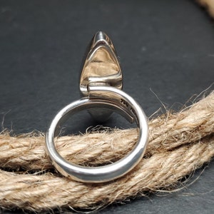 May include: A silver ring with a large, oval-shaped stone. The ring band is smooth and rounded, with a split design near the stone. The stone is a light color, and the ring is set against a neutral background.