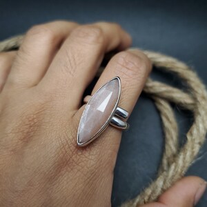 May include: A silver ring featuring a large, elongated, faceted, light peach-colored gemstone. The ring has a double-band design and is shown on a person's finger. The gemstone is set in a silver bezel.