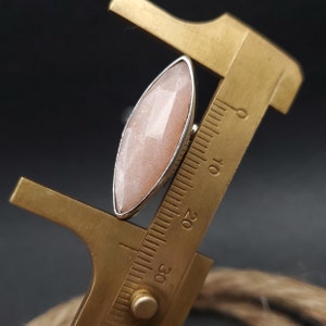 May include: A silver ring with a large, elongated, peach-colored gemstone. The gemstone has a faceted surface. The ring is being measured with a brass caliper. The background is dark.