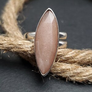 May include: A silver ring featuring a large, elongated, faceted, peach-colored gemstone. The gemstone is set in a silver bezel and has a pointed, leaf-like shape. The ring band is split into two parallel silver bands.
