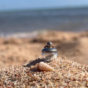 Könnte beinhalten: Ein kleines Glasgefäß mit silbernem Deckel, gefüllt mit weißem Sand, liegt an einem Sandstrand. Eine kleine Muschel liegt neben dem Glas. Der Hintergrund zeigt das Meer und einen blauen Himmel.