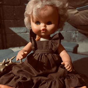 May include: A vintage doll with blue eyes and blonde, curly hair. The doll wears a brown dress with ruffled details. The doll is sitting with its arms and legs extended. The doll's skin is a light peach color.