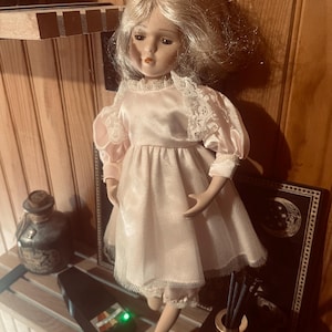 Peut inclure: Une poupée en porcelaine aux cheveux blonds, vêtue d'une robe en satin rose pâle avec des manches bouffantes et une bordure en dentelle blanche. La poupée présente des traits peints, dont des lèvres rouges et des yeux sombres, et une jupe en tulle.