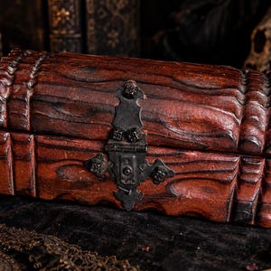 Può includere: Un forziere rettangolare in legno con finitura marrone rossastro scuro e coperchio curvo. La scatola presenta un vistoso meccanismo di chiusura in metallo nero usurato centrato sul davanti, con profonde scanalature orizzontali intagliate.