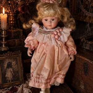 May include: A porcelain doll with blonde curly hair styled with a pink bow, wearing a peach-colored satin dress featuring a ruffled collar with lace trim and small pink decorative spheres, paired with light-colored shoes on its feet.