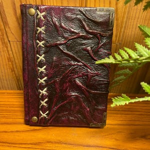 Peut inclure: Un journal en cuir violet foncé avec un design cousu décoratif et des accents en laiton. Le journal a un aspect texturé et vieilli.