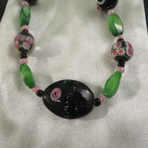 May include: A black and green glass bead necklace with pink floral accents. The necklace features a large oval bead with a pink flower design.