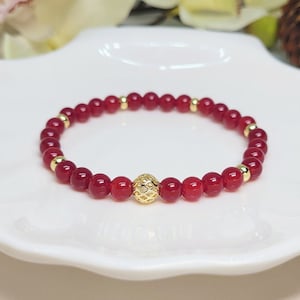 May include: A red gemstone bracelet with a gold bead charm. The bracelet is made of small, round beads and is strung on a clear elastic cord.