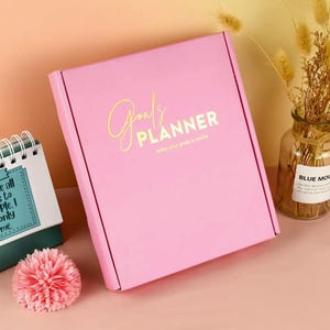May include: A pink Goals Planner with gold lettering and the tagline "make your goals a reality." The planner is rectangular and closed, with a small calendar and a pink flower in the foreground. A glass jar with dried flowers is in the background.