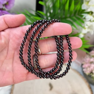 May include: A strand of dark red garnet beads, strung on a clear cord, is held in a hand. The beads are small and round, and the strand is about 18 inches long.