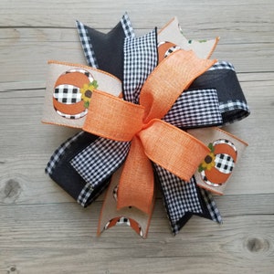 Op de afbeelding: Een grote, meerlagige strik gemaakt van zwart-wit buffalo check lint, oranje lint en jute lint met een pompoenontwerp. De strik is vastgebonden met een groot oranje lint in het midden.