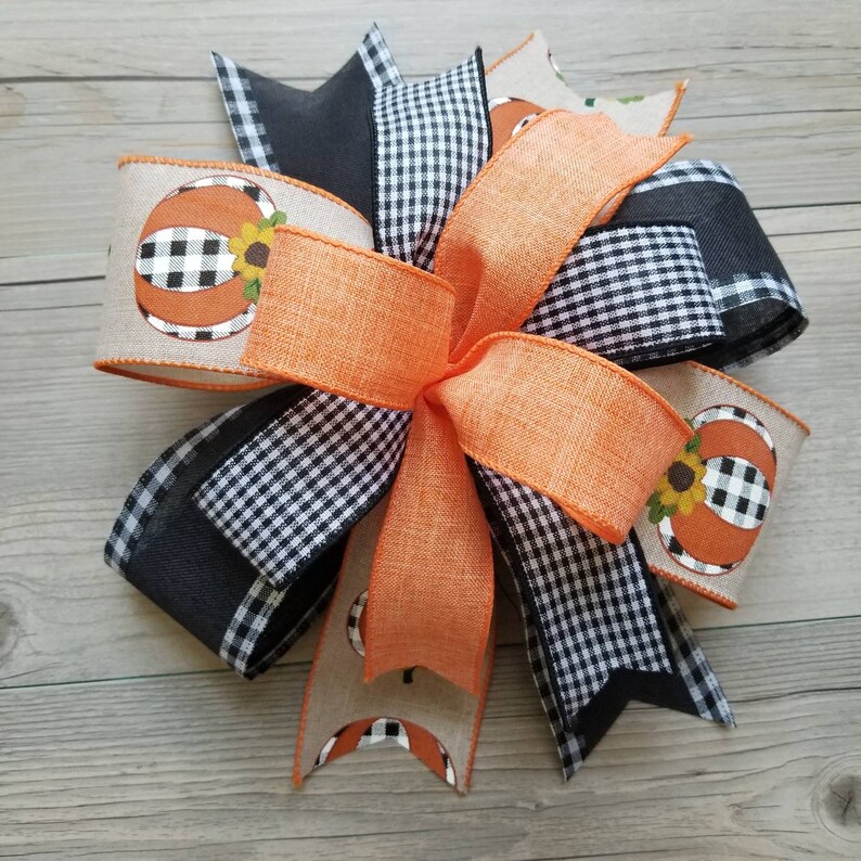 Op de afbeelding: Een grote, oranje en zwarte geruite strik met een jute lint met een pompoenontwerp. De strik is vastgebonden met een zwart lint en heeft een zonnebloem op de pompoen.