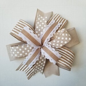 May include: A decorative bow crafted from multiple ribbons featuring a mix of tan and white patterns. The design includes solid tan linen-textured ribbon, white polka dots on a tan background, white stripes on a tan background, and white lace trim.