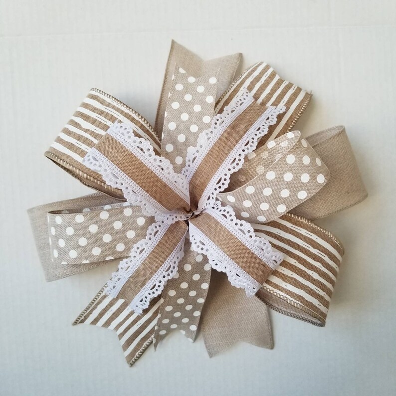 May include: A decorative bow crafted from multiple ribbons featuring a mix of tan and white patterns. The design includes solid tan linen-textured ribbon, white polka dots on a tan background, white stripes on a tan background, and white lace trim.