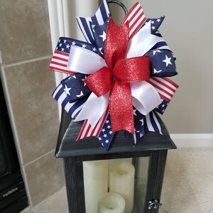 Op de afbeelding: Een decoratieve zwarte lantaarn met drie witte kaarsen erin, bekroond met een patriottische strik. De strik heeft rode, witte en blauwe linten met sterren en strepen, wat een feestelijke uitstraling cre&euml;ert.