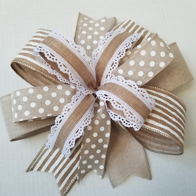 May include: A decorative bow crafted from tan and white ribbon featuring a mix of polka dot and striped patterns. The center of the bow is accented with a delicate white lace trim that edges the loops and tails, creating a layered appearance.