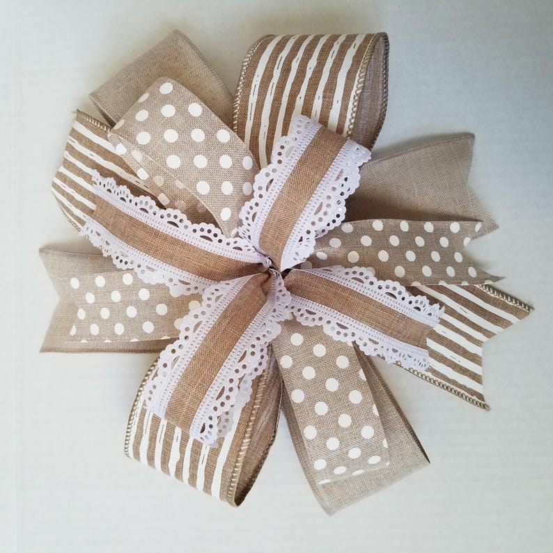 May include: A decorative bow crafted from multiple ribbons featuring a mix of solid tan, white polka dot, and white vertical striped patterns. The center of the bow is accented with a white lace trim that features a scalloped edge design.