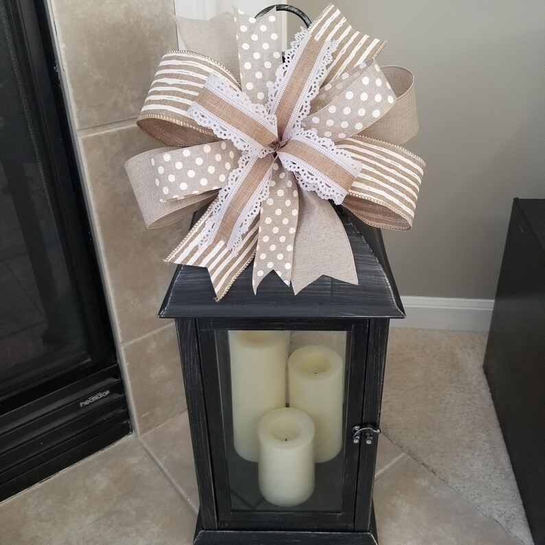 May include: A black lantern with a distressed finish containing three cream-colored pillar candles. A large decorative bow sits on top, featuring a mix of tan, white, and beige ribbons with polka dot, striped, and solid patterns, plus white lace trim.
