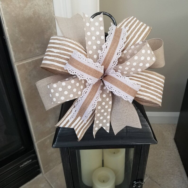 May include: A decorative bow featuring a mix of tan and white ribbons with polka dot and horizontal stripe patterns. The bow includes a central layer of solid tan ribbon edged with white lace trim, all arranged in a multi-loop design.